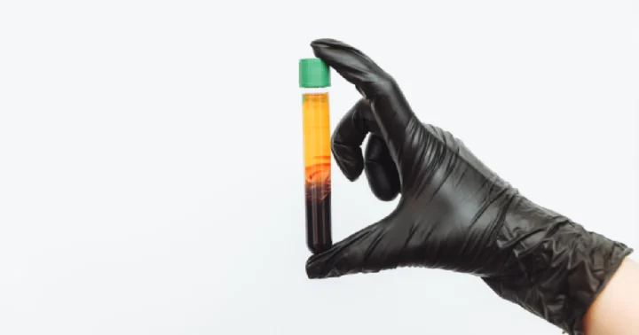 A hand in a black glove holds a test tube with a green cap, showing a liquid separated into yellowish-orange and reddish-brown layers at Ohh Shot in Kenai, AK.