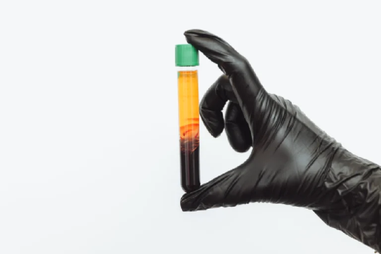 A hand in a black glove holds a test tube with a green cap, showing a liquid separated into yellowish-orange and reddish-brown layers at Ohh Shot in Kenai, AK.