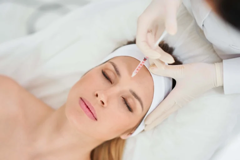 Young woman receives a forehead injection from a gloved professional, lying back with a white headband and towel during Botox and Dysport Treatments in Kenai, AK.
