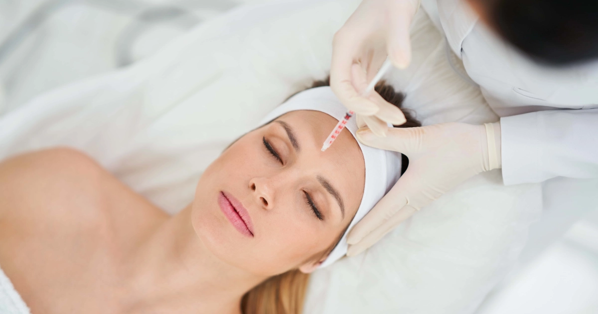 Young woman receives a forehead injection from a gloved professional, lying back with a white headband and towel during Botox and Dysport Treatments in Kenai, AK.
